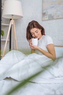 Yatakta çay tutan mutlu genç bir kadının seçici odak noktası. 