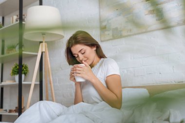 Yatakta çay tutan neşeli genç bir kadının seçici odağı. 