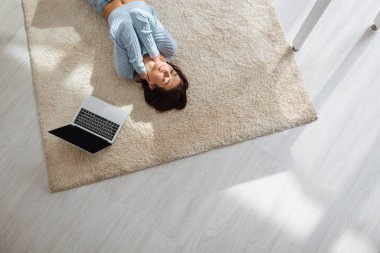 top view of happy woman resting while lying on carpet near laptop at home  clipart