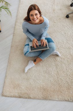 Halıda otururken dizüstü bilgisayar kullanan neşeli blog yazarının üst görünümü 