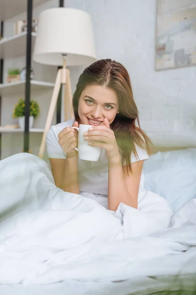 快乐的年轻女子一边在床上端着茶杯一边看着相机的选择性焦点 — 图库照片