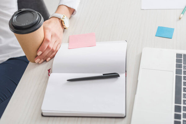 обрезанный вид деловой женщины с бумажной чашкой возле ноутбука и ноутбука в офисе
 