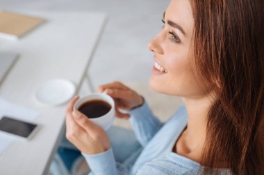 Evde hayal kurarken elinde kahveyle gülümseyen bir kadının seçici odağı. 