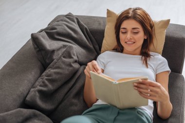 Oturma odasındaki kanepede dinlenirken kitap okuyan neşeli genç bir kadın. 