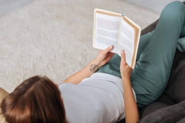 Oturma odasındaki kanepede dinlenirken kitap okuyan genç bir kadının genel görünümü. 