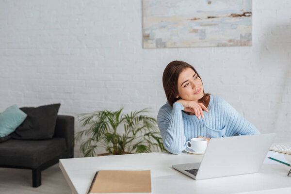 dreamy woman looking away near laptop and cup on table 