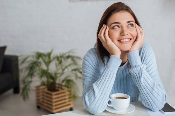 dreamy young woman smiling near cup with coffee 