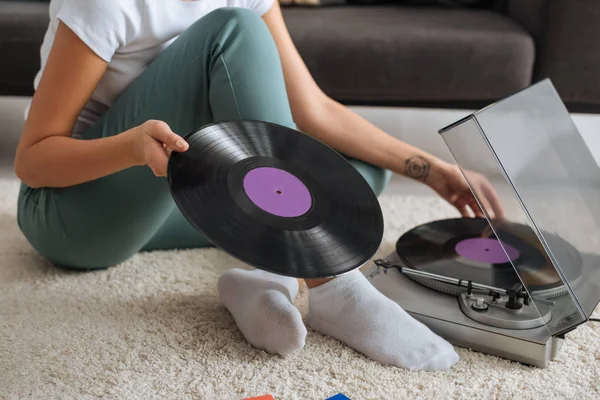 カーペットの上に座ってレトロなレコードプレーヤーの近くにビニールレコードに触れるタトゥーの女の子のクロップドビュー — ストック写真