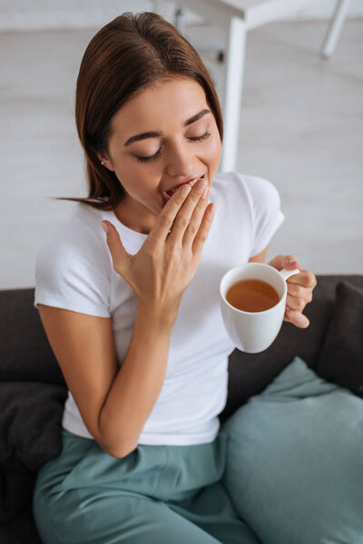 overhead view of tired woman yawning and cover mouth while holding cup of tea 