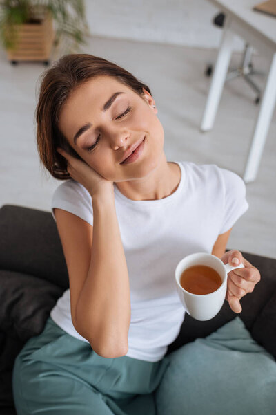 young dreamy woman holding cup with tea while chilling on sofa in living room 
