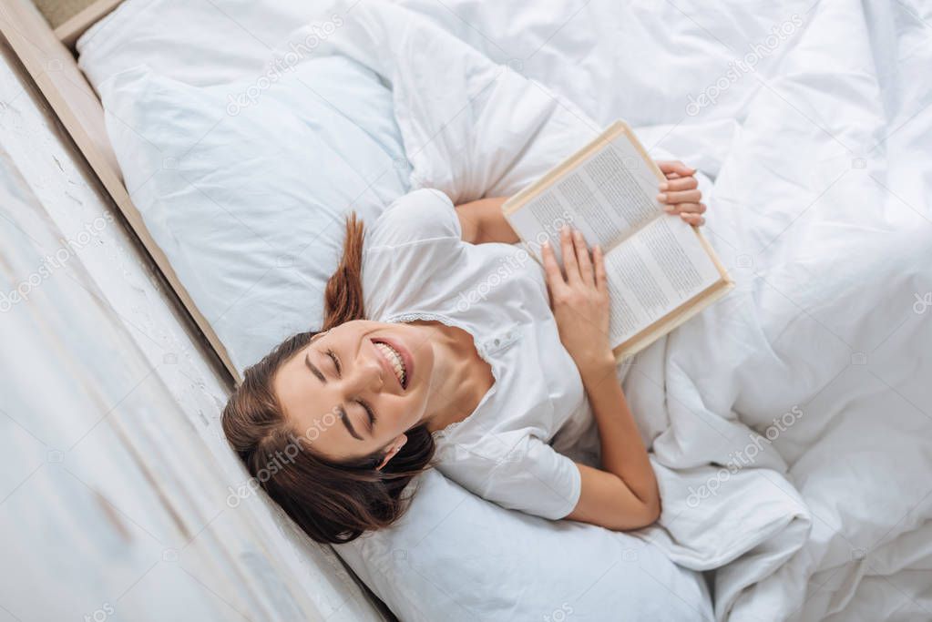 top view of happy girl holding book while chilling in bed 