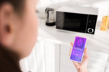 selective focus of woman holding smartphone with shopping app and microwave on background 