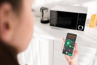selective focus of woman holding smartphone with marketing analysis app and microwave on background 