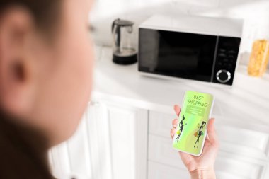 selective focus of woman holding smartphone with best shopping app and microwave on background 