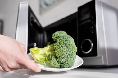 cropped view of woman holding plate with broccoli near microwave 