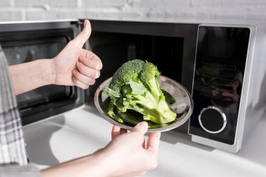 cropped view of woman holding plate with broccoli near microwave and showing like  clipart