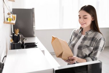 smiling woman holding paper bag with popcorn near microwave in kitchen 