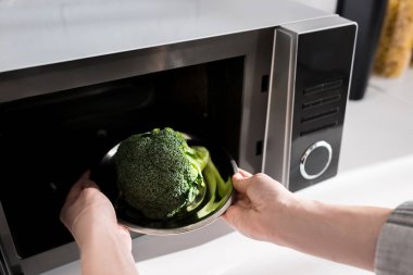 cropped view of woman holding plate with broccoli and putting it in microwave  clipart