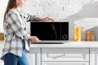 cropped view of smiling woman in shirt looking at microwave in kitchen 