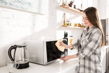 smiling and attractive woman cleaning microwave with rag in kitchen 