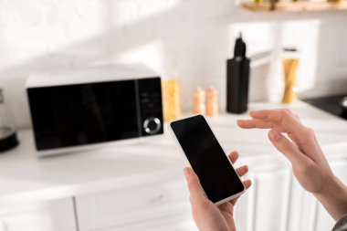 cropped view of woman using smartphone near microwave in kitchen 