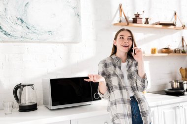 smiling woman talking on smartphone near microwave in kitchen 