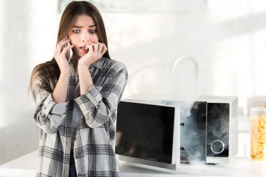 Mutfakta bozuk mikrodalga fırının yanında akıllı telefondan konuşan korkmuş bir kadın. 