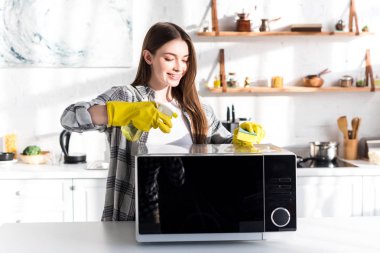 smiling woman in rubber gloves cleaning microwave with sponge