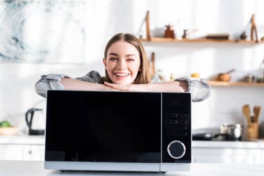 smiling and attractive woman standing near microwave in kitchen 