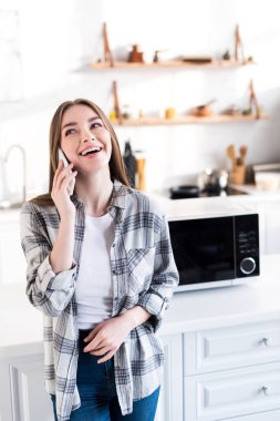 smiling woman talking on smartphone near microwave in kitchen 