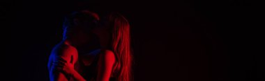 Siyah panoramik çekimde izole edilmiş kırmızı ışıkta öpüşen ve sarılan seksi çiftin yan görüntüsü.