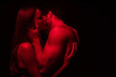Kırmızı ışıkta öpüşen, tutkulu, çıplak ve seksi genç çiftin yan görünüşü siyahta izole.