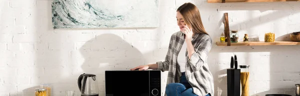 Plano Panorámico Mujer Sonriente Hablando Teléfono Inteligente Cerca Microondas Cocina —  Fotos de Stock