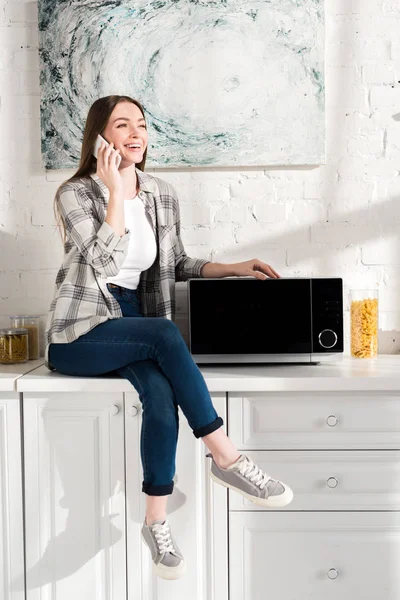 Mujer Sonriente Hablando Teléfono Inteligente Cerca Microondas Cocina — Foto de Stock