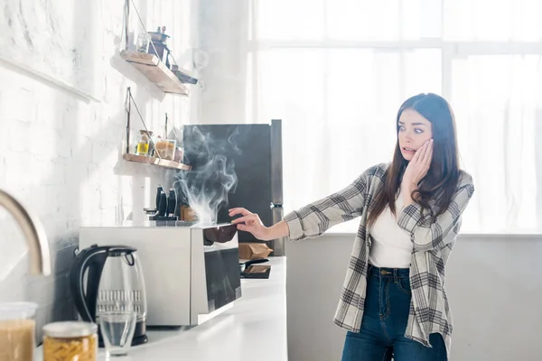 scared and attractive woman looking at broken microwave in kitchen