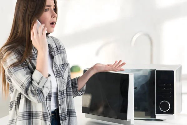 Shocked Woman Talking Smartphone Broken Microwave Kitchen — Stock Photo, Image