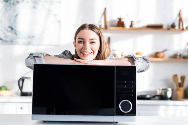 улыбающаяся и привлекательная женщина стоит рядом с микроволновой печью на кухне
 