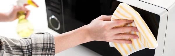 panoramic shot of woman cleaning microwave with rag in kitchen