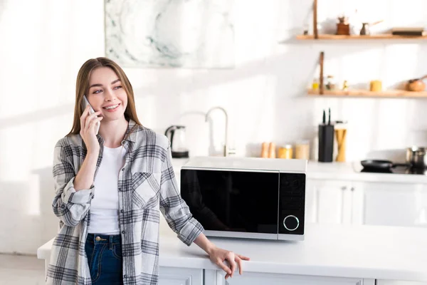 Mujer Sonriente Hablando Teléfono Inteligente Cerca Microondas Cocina — Foto de Stock