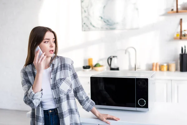 Mujer Conmocionada Hablando Teléfono Inteligente Cerca Microondas Cocina — Foto de Stock