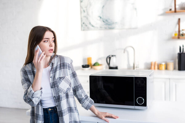 шокированная женщина разговаривает на смартфоне рядом с микроволновой печью на кухне
 