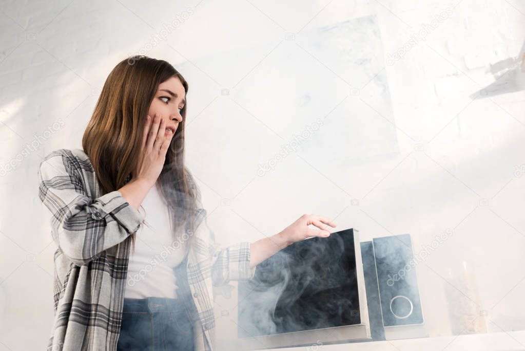 low angle view of shocked and attractive woman looking at broken microwave in kitchen 