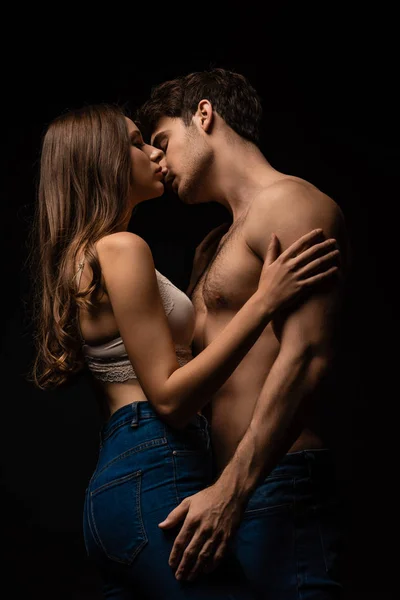 穿着牛仔裤拥抱和亲吻隔离在黑人身上的性感年轻夫妇的侧面视图 — 图库照片