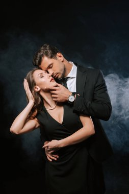 Şehvetli bir adam siyah elbiseli çekici bir kadına dumanla dokunuyor. 