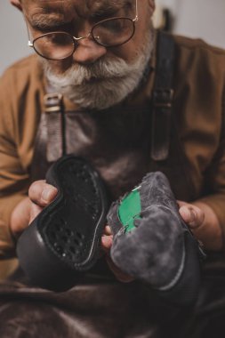 senior, bearded cobbler holding shoe and sole in workshop clipart