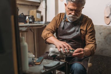 selective focus of senior, bearded shoemaker repairing shoe in workshop clipart