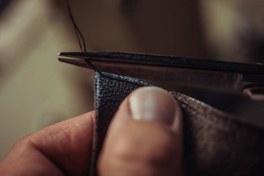 cropped view of cobbler cutting thread near piece of genuine leather clipart