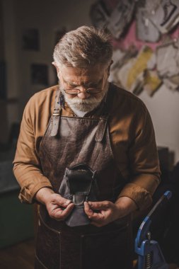 attentive, senior cobbler holding leather detail in workshop clipart