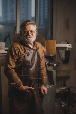 positive, bearded shoemaker in leather apron looking at camera while standing in workshop clipart