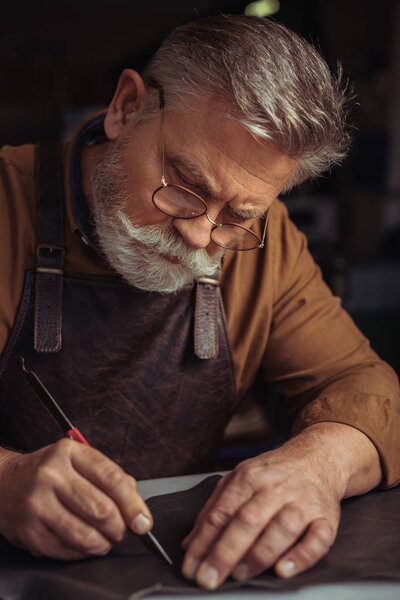 senior, bearded shoemaker cutting genuine leather with knife